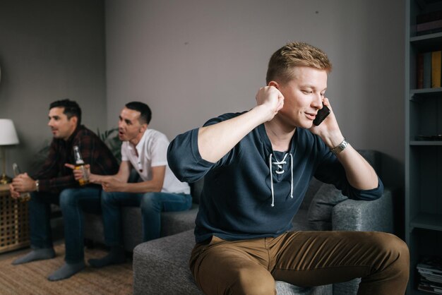 Beau mec essayant de parler au téléphone Derrière des amis regardent la télévision Groupe d'amis regardant un match de football à la télévision sur fond de conversation téléphonique d'un jeune homme