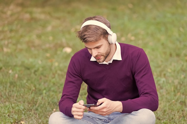 Beau mec sur l'herbe verte change de chanson sur le lecteur mp3 du téléphone portable et écoute de la musique dans les écouteurs à l'ère numérique