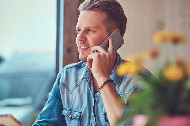 Beau mec hipster avec une coupe de cheveux et une barbe élégantes est assis à une table dans un café en bordure de route, parlant au téléphone.