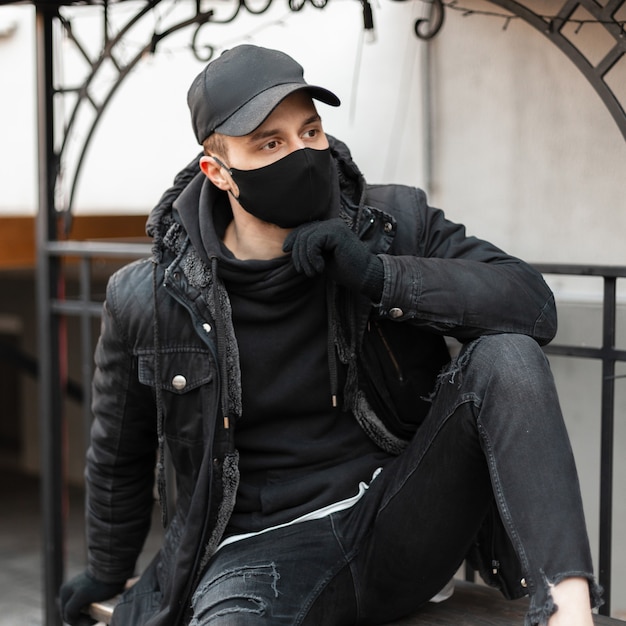 Beau mec hipster à la mode avec une casquette élégante noire et un masque médical de protection dans une veste noire et un sweat à capuche se trouve dans la ville. Style moderne décontracté masculin urbain