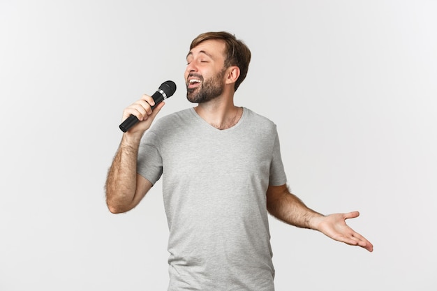 Beau mec insouciant chantant la chanson au karaoké, tenant le microphone, debout sur fond blanc