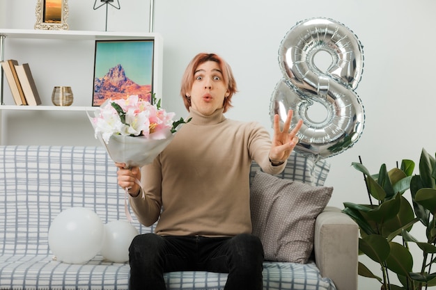 Beau mec le jour de la femme heureuse tenant un bouquet assis sur un canapé dans le salon