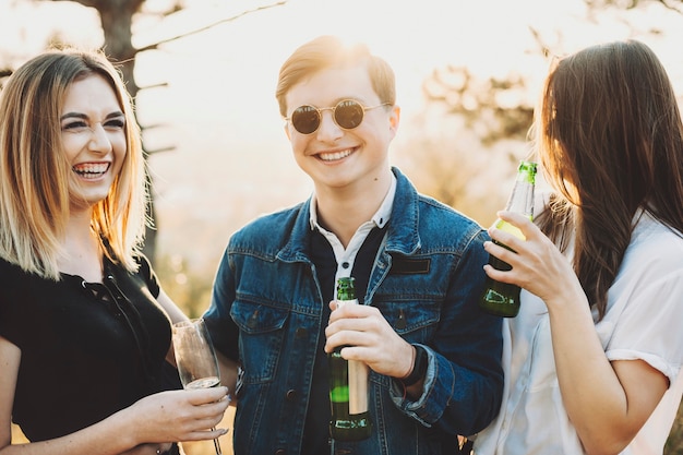 Beau Mec En Lunettes De Soleil élégantes Souriant Et Tenant Une Bouteille De Bière Tout En Passant Du Temps Dans La Nature Avec Deux Jeunes Femmes. Homme Gai, Boire De L'alcool Avec Des Amies