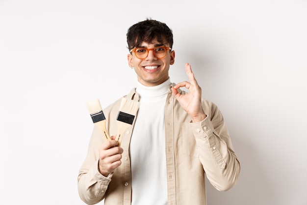 Beau mec naturel dans des verres recommandant le magasin, montrant des pinceaux pour la rénovation et la décoration, signe d'approbation, debout sur un mur blanc.