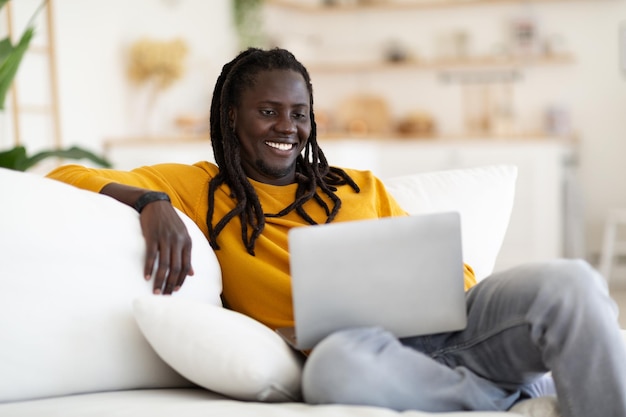 Beau mec noir avec des dreadlocks relaxant avec un ordinateur portable sur un canapé à la maison