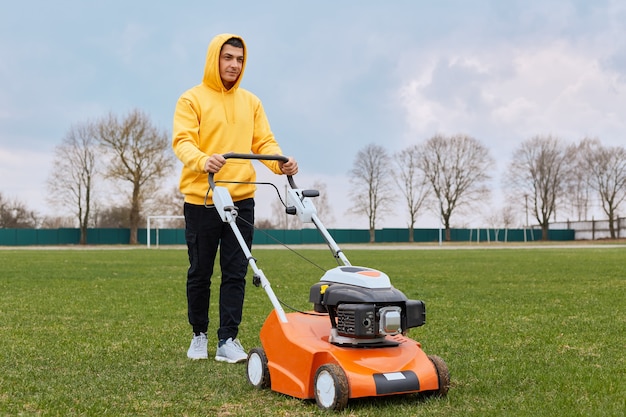 Beau mec pensif en sweat à capuche jaune et pantalon noir avec tondeuse à essence dans ses mains tond l'herbe sur la pelouse