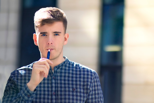 Beau mec sérieux, jeune homme réfléchi fume, produit de tabac de chauffage, système à l'extérieur. Garçon avec une nouvelle e-ciggarette à la main dans le smog, la fumée, prend une bouffée. Alternative au tabagisme. Mauvaise habitude.