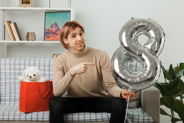 Beau mec souriant le jour de la femme heureuse tenant et pointant le ballon numéro huit assis sur un canapé dans le salon