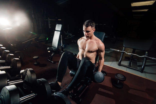 Beau mec torse nu soulevant des haltères lourds alors qu'il était assis sur un banc dans une salle de sport sombre