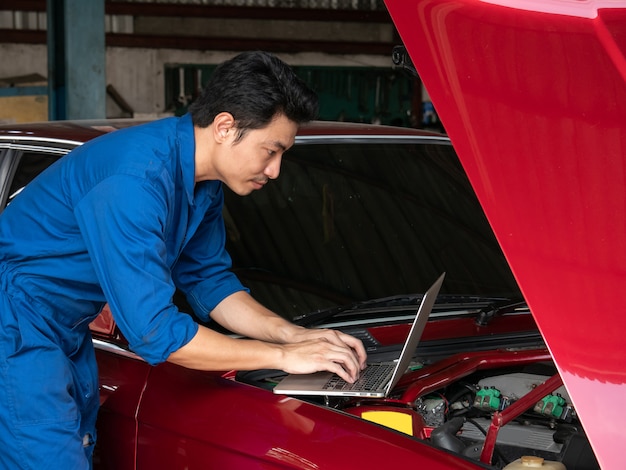 Beau mécanicien asiatique utilisant un ordinateur portable pour vérifier un moteur de voiture et montrant les pouces au garage de réparation.