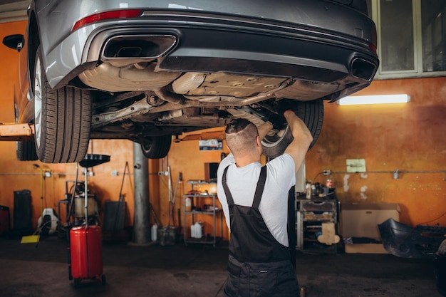 Un beau mécanicien automobile professionnel enquête sur la rouille sous un véhicule sur un ascenseur en service Un réparateur utilise une lampe LED et se dirige vers un spécialiste portant des lunettes de sécurité Atelier moderne