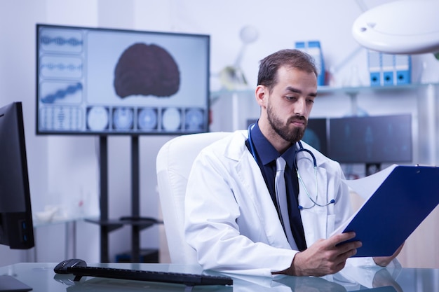 Beau médecin barbu vérifiant la liste des patients à visiter pour la journée. Moniteur avec cerveau 3d en arrière-plan.