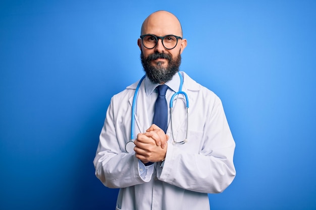 Beau médecin chauve avec barbe portant des lunettes et stéthoscope sur fond bleu avec les mains jointes et les doigts croisés souriant détendu et joyeux Succès et optimiste