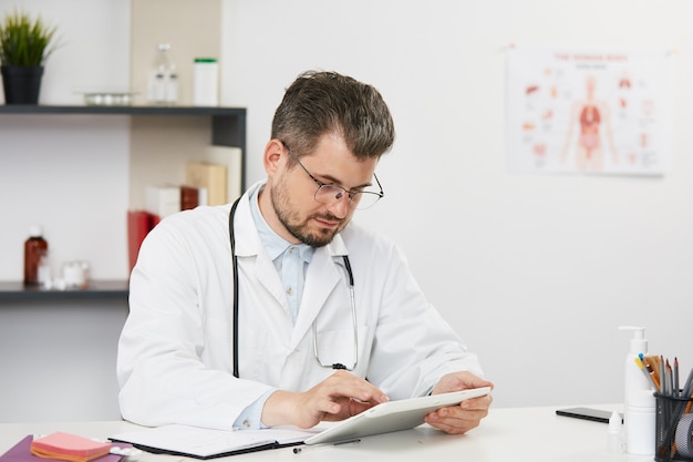 Beau médecin senior à l'aide de tablette dans son cabinet médical, thérapeute masculin barbu en uniforme médical blanc et lunettes assis dans le cabinet médical avec stéthoscope et travaillant sur tablette