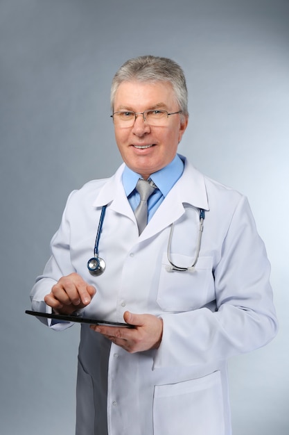 Un beau médecin avec stéthoscope et tablette debout sur fond gris