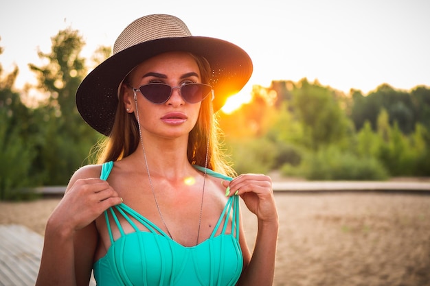 Un beau modèle brune dans un maillot de bain vert lunettes noires et un chapeau pose devant la caméra