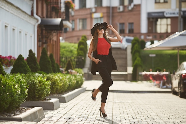 Beau modèle brune femme avec un corps parfait dans le chemisier rouge et dans le costume noir à la mode en ajustant son élégant chapeau noir, sourit et posant dans la rue ensoleillée de la ville