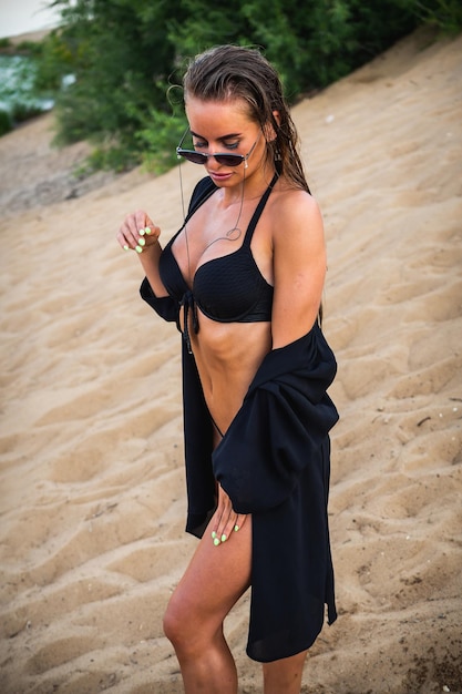 Un beau modèle brune en maillot de bain noir et lunettes noires posant sur le sable devant la caméra