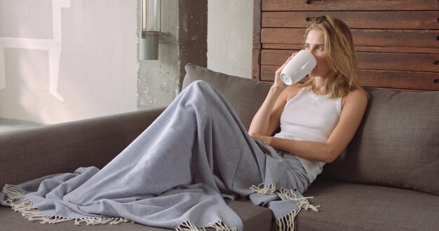 Beau modèle féminin sur un canapé avec une tasse de café sous une couverture bleue en regardant la caméra puis en revenant à ses pensées