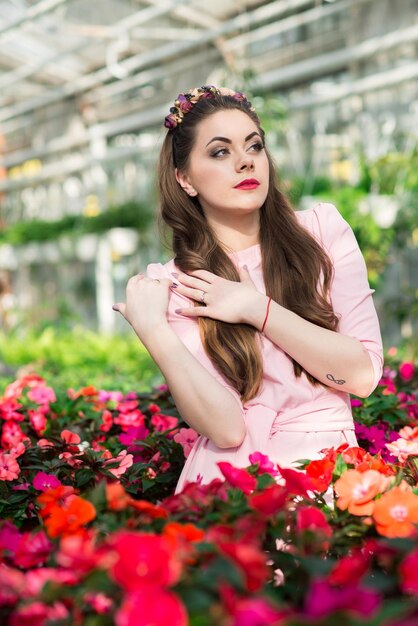 Beau modèle féminin vêtu d'une longue robe rose pose parmi les nombreuses fleurs de la serre