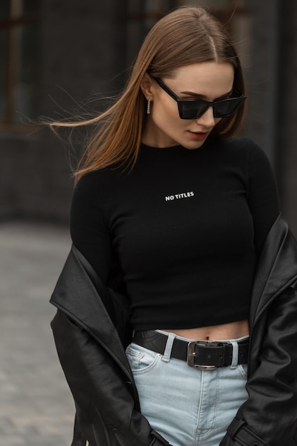 Beau modèle de femme hipster à la mode avec des lunettes de soleil cool dans un long manteau en cuir à la mode, un t-shirt noir et un jean taille haute se promène dans la rue