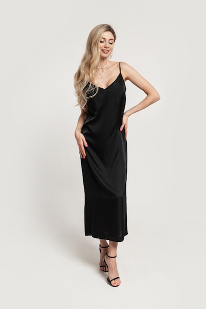 Beau modèle de femme sexy de mode élégante aux cheveux blonds dans une robe noire à la mode à la mode avec des chaussures à talons posant sur un fond blanc dans le studio