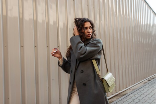 Beau modèle de fille bouclée avec un long manteau vert à la mode avec un sac marche dans la rue près d'un mur en métal