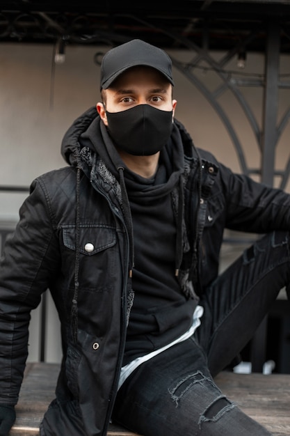 Beau modèle de jeune homme avec casquette noire et masque médical de protection en veste noire et sweat à capuche se trouve dans la rue