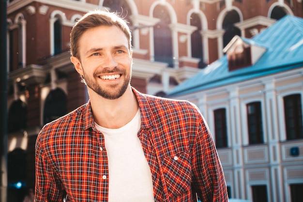 Beau modèle lambersexuel hipster élégant et souriant. Homme moderne vêtu d'une chemise à carreaux rouge. Homme de mode posant près de gratte-ciel sur le fond de la rue en lunettes de soleil. À l'extérieur au coucher du soleil