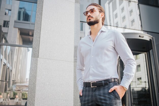 Beau modèle masculin se tient debout et pose. Il a l'air élégant. Jeune homme porte des lunettes de soleil et attend avec impatience. Il est attrayant et beau.