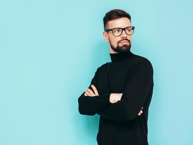 Beau modèle souriant Sexy homme élégant vêtu d'un pull à col roulé noir et d'un jean Mode hipster mâle posant près du mur bleu en studio Isolé dans des lunettes ou des lunettes Bras croisés