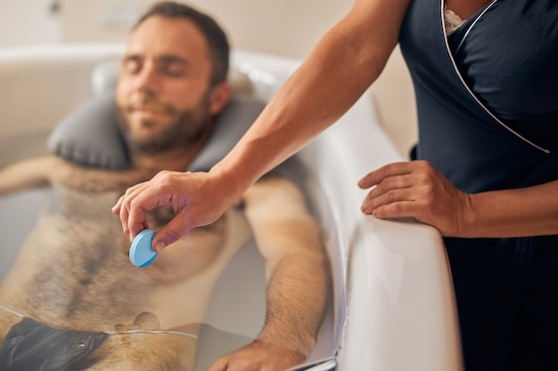 Beau monsieur allongé dans la baignoire tandis que l'esthéticienne met la tablette de bain relaxante dans l'eau