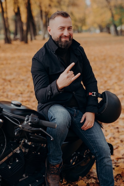 Beau motard barbu fait des gestes de corne avec les doigts, se sent cool, porte un manteau noir et un jean, s'assoit sur une moto rapide