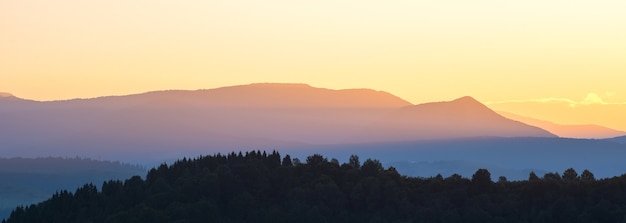 Beau panorama montagneux de paysage avec des pics brumeux et une vallée boisée brumeuse au coucher du soleil.