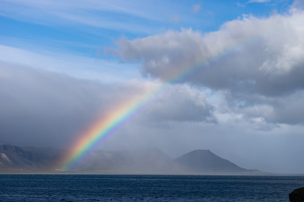 Beau paysage d'été avec mer, petit village sur la colline, arc-en-ciel coloré lumineux sur ciel bleu, nuages et montagnes à l'horizon