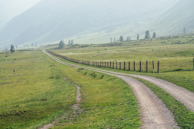 Beau paysage de montagne verte avec un long chemin de terre le long d'une clôture et de grandes montagnes dans le brouillard. Paysage de montagne brumeux atmosphérique avec chemin de terre parmi les collines et les grandes montagnes. Longueur route en campagne