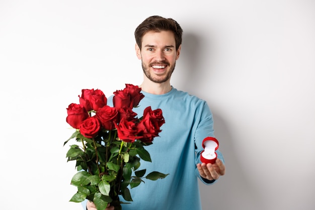 Beau petit ami de jeune homme faisant une proposition le jour des amoureux de la Saint-Valentin, tenant un bouquet de roses rouges et une bague de fiançailles, concept de mariage et de relation.