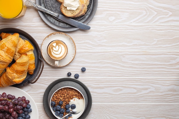 Photo beau petit-déjeuner matinal avec croissants frais, granola avec yaourt et baies, toasts, cappuccino de café et fruits sur assiette. petit-déjeuner savoureux et sain sur une table en bois blanc, espace pour le texte