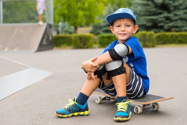 Beau petit garçon assis sur sa planche à roulettes dans son équipement de sécurité souriant à la caméra alors qu'il fait une pause au skate park