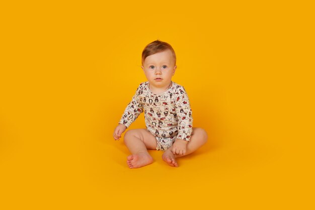 un beau petit garçon aux yeux bleus dans un body est assis sur un fond jaune