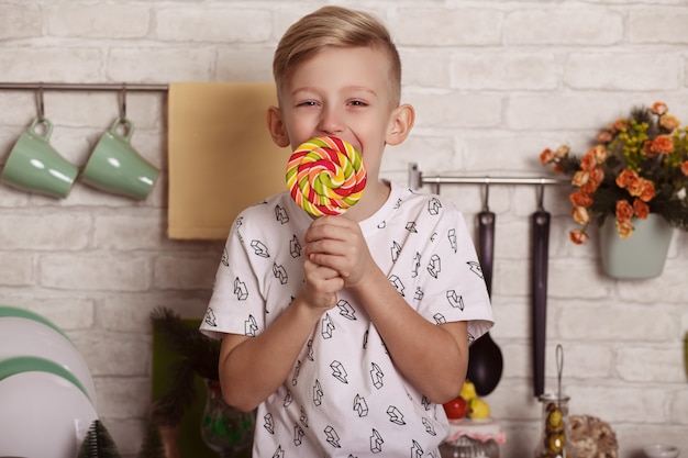 Beau petit garçon blond est assis sur la table de la cuisine avec une grosse sucette à la main