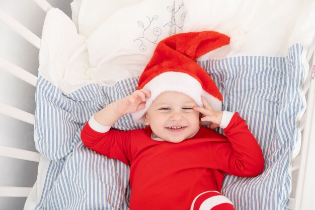 Beau Petit Garçon En Bonnet De Noel, Costume Rouge Souriant Dans Un Lit Bébé à La Maison.