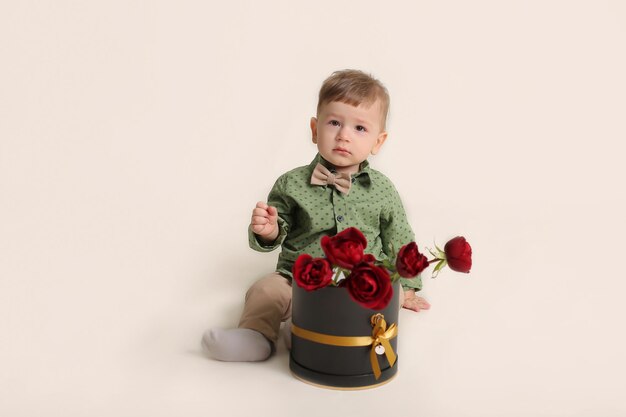 un beau petit garçon dans une chemise verte est assis sur un fond blanc à côté d'une boîte avec des roses rouges