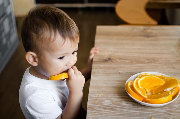 Beau Petit Garçon Dans La Cuisine Mangeant Avec Impatience Une Orange, Coupée En Quartiers