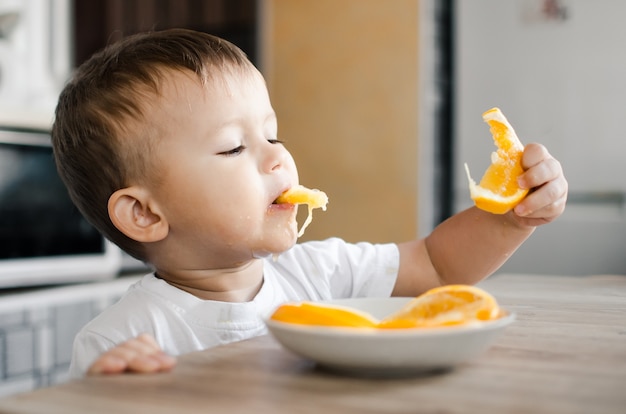 Beau petit garçon dans la cuisine mangeant avec impatience une orange, coupée en quartiers