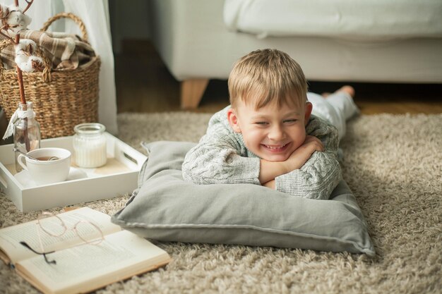 Un beau petit garçon dans un pull tricoté lit près de la fenêtre avec une tasse de thé chaud. Confortable. L'automne. Humeur d'automne.