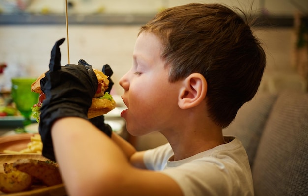 Le beau petit garçon mangeant un hamburger dans des gants en caoutchouc noir