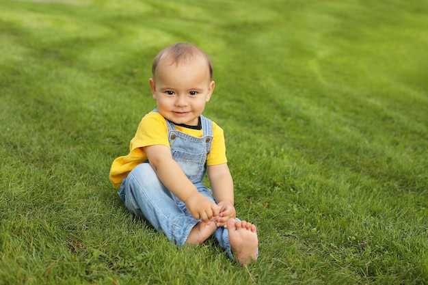 un beau petit garçon vêtu d'un t-shirt jaune et d'un costume en jean est assis sur la pelouse du parc