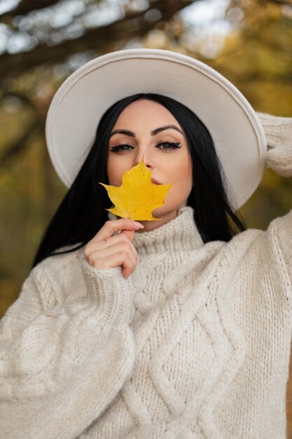 Beau portrait d'automne d'une belle jeune fille dans un pull vintage à la mode et un chapeau couvrant son visage avec une feuille d'automne jaune dans le parc