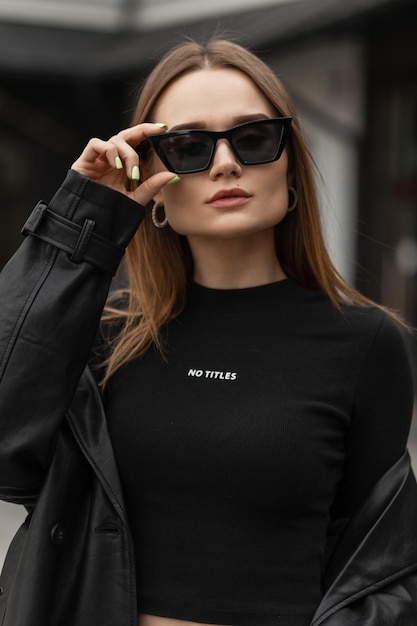 Beau portrait urbain féminin d'une belle jeune fille modèle dans des vêtements noirs à la mode porte des lunettes de soleil de mode cool noires dans la rue